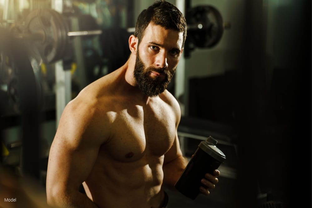 Side view portrait of young muscular man bodybuilder shirtless sitting in dark gym holding protein supplement shaker in training waist up black hair and beard looking to the camera